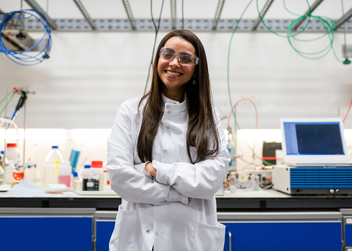woman in white coat and safety googles smiling in a lab