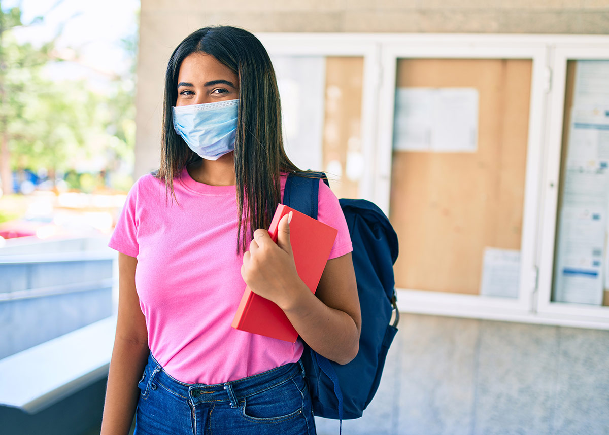 woman on a university campus wearing a facemask and backpack and holding a book
