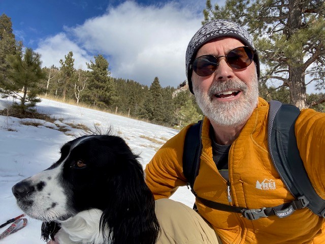 Man wearing an orange coat, glasses, and a hat smiling in a selfie with a dog